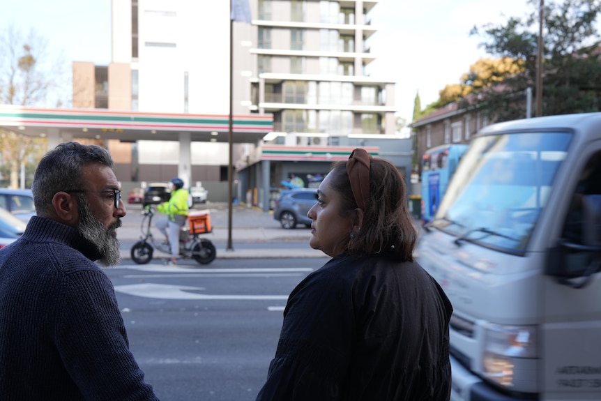 A bearded man and dark-haired woman stand across the street from a 7-Eleven petrol station.