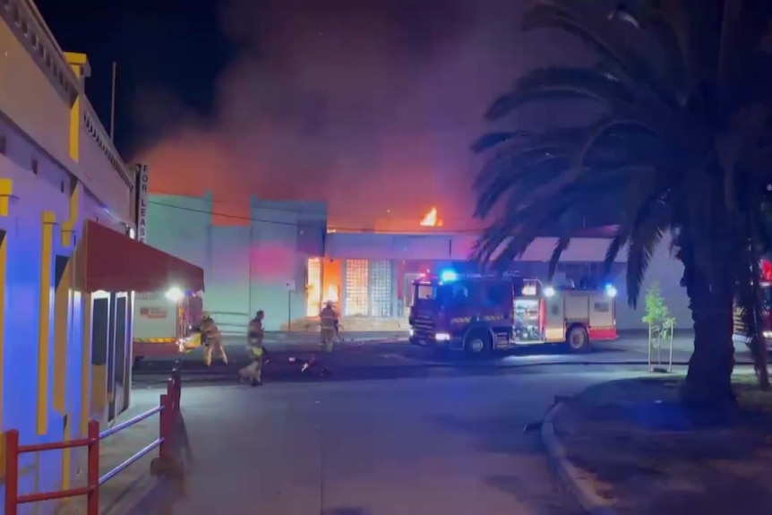 Three firefighters outside a building which has orange flames coming from the roof at night.