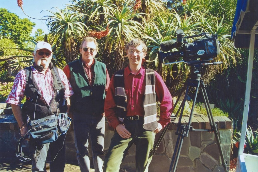 A young Jerry Coleby-Williams stands next to a film camera and two cameramen in a garden