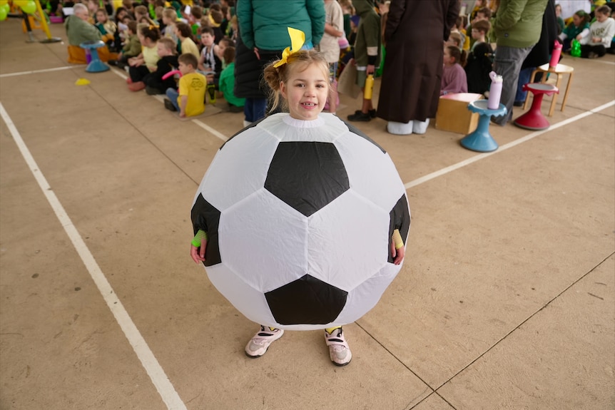 A young girl stands outside on a basketball court dressed in an inflatable soccer ball with a gold ribbon in her hair.