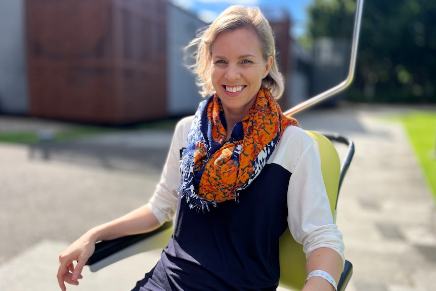 A blonde-haired woman smiles widely as she sits on a chair in an outdoor area