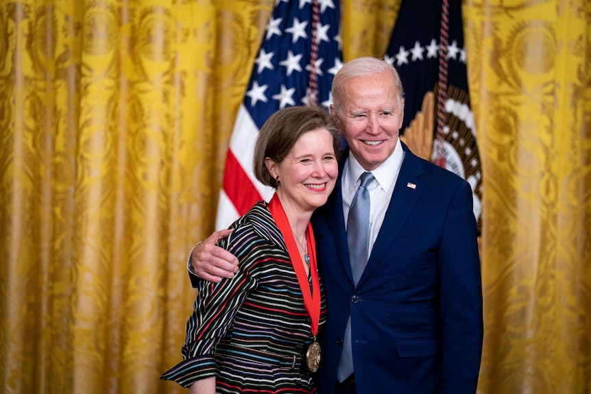 President Joe Biden with his arm around Ann Patchett, who has a medal around her neck, the US flag behind them.