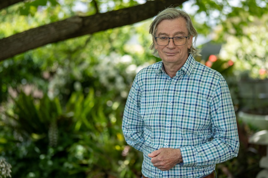 A man in a checkered green shirt and glasses stand under a tree in a garden