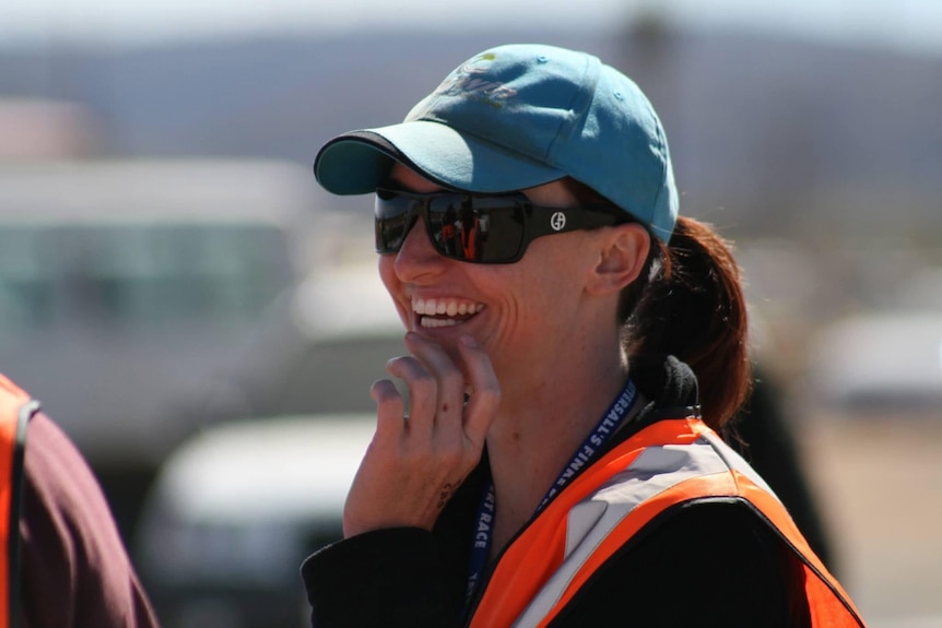 A woman, wearing a cap and a high-viz vest, smiling.