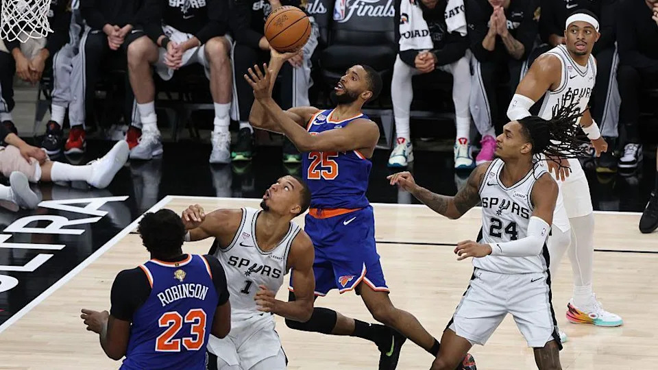 A Knicks player leaps towards the basket with the ball in hand as opposing Spurs players surround him
