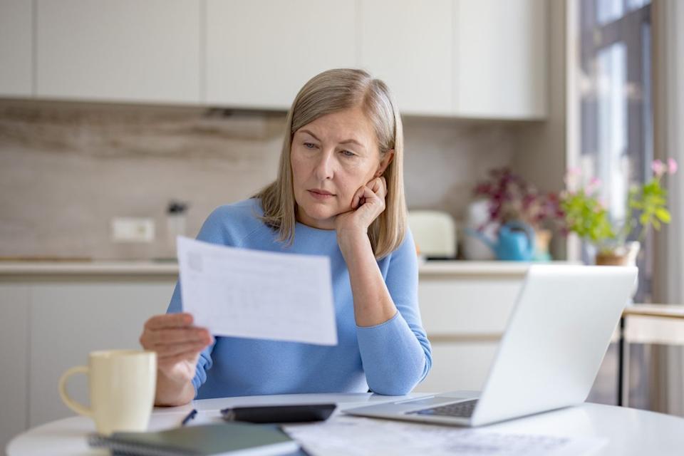 An older woman with light hair wearing a blue sweater sits at a white table, intently reading documents. Her left hand rests on her chin. A silver laptop, a yellow mug, a calculator, and notebooks are also on the table. The background shows a modern kitchen or dining area with white cabinets and a window.