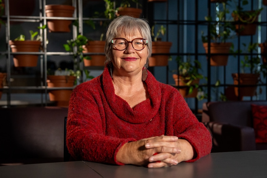 A woman with greying hair sits with her fingers interlaced on the table in front of a rack full of indoor plants