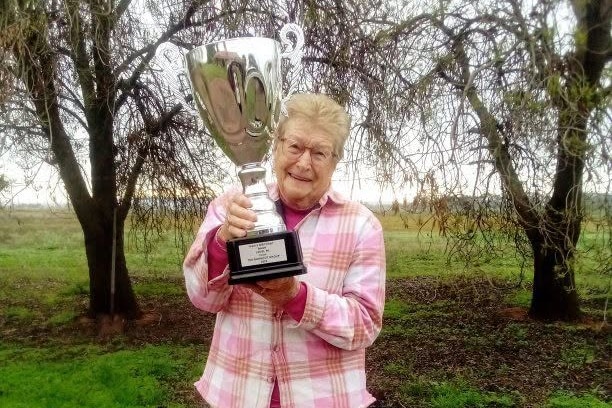 An older woman with glasses and blonde hair with a pink jacket stands smiling with a big silver trophy