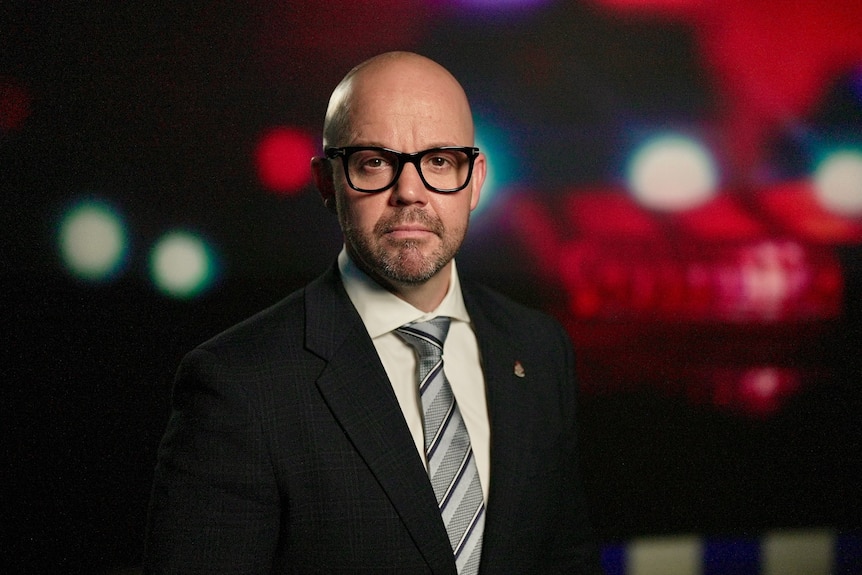 A man in a suit and thick-rimmed black glasses looking at the camera in front of police-lights background.