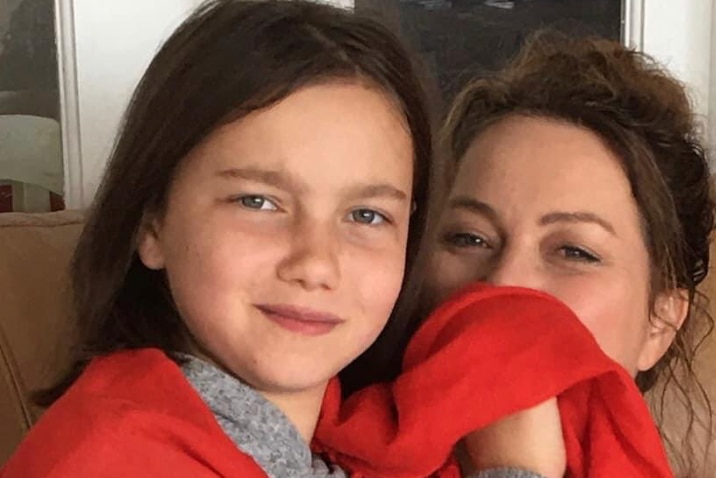 A smiling, dark-haired girl cuddles up to her mum, whose face is partially hidden behind a blanket.