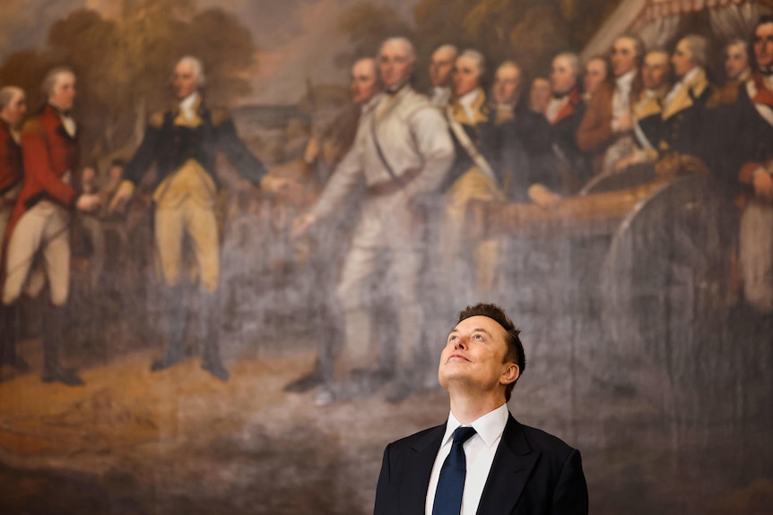 Elon Musk looks up into the air, with a huge portrait behind him