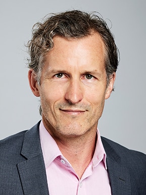 A dark-haired middle aged man in a dark blazer smiles for a corporate headshot.