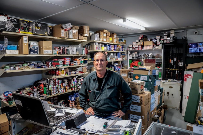 A man in a green shirt sits in a room at a desk. There is a laptop in front of him and shelves with bullets behind.