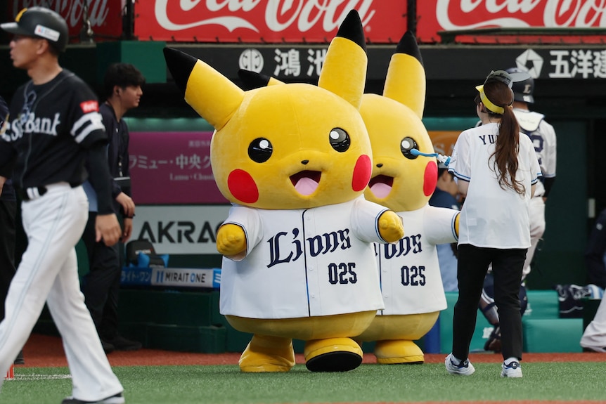 Two Pikachu mascots on a baseball field as a player walks past.