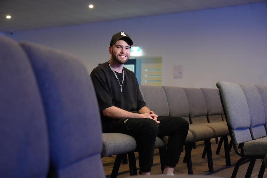 Jake smiles as he sits on a chair in an auditorium wearing black clothing
