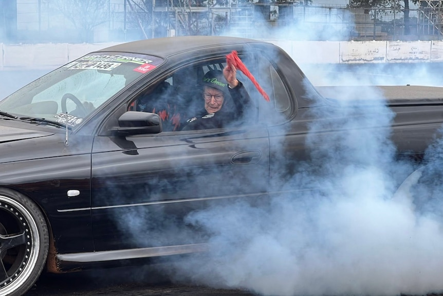 A woman with a helmet on waves an orange flag out a black ute with grey smoke coming out the back.