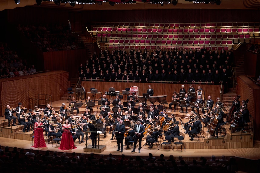 Simone Young leads a performance with full orchestra and a large choir before a seated audience in a concert hall.