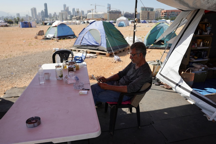A man sitting on a chair outside a tent in a field with other tents in the background.