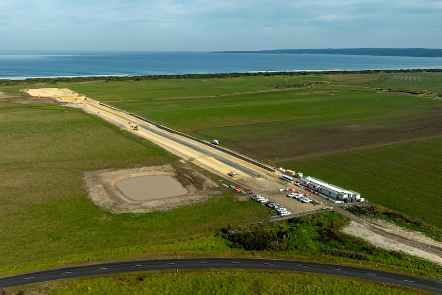 An aerial photo of a large strip of dirt and dozens of cars and a shed-like building a few hundred metres from a beach.