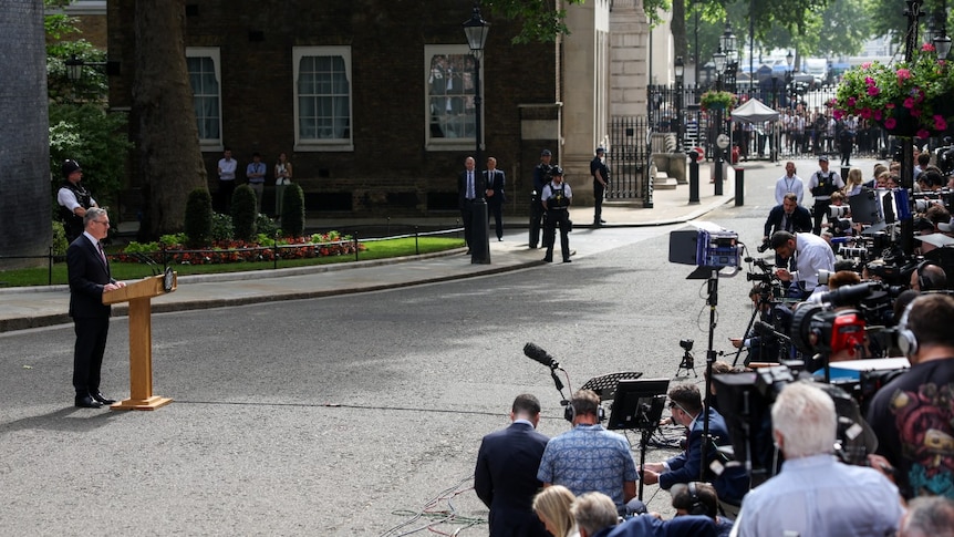 Keir Starmer, wearing a suit, speaks at a lectern while a large number of media professionals look on.