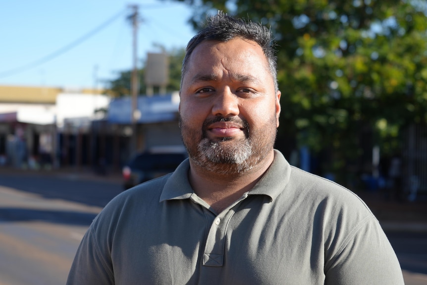 Chest-up shot of Indian-Australian man with black hair and beard, standing street-side with serious expression.