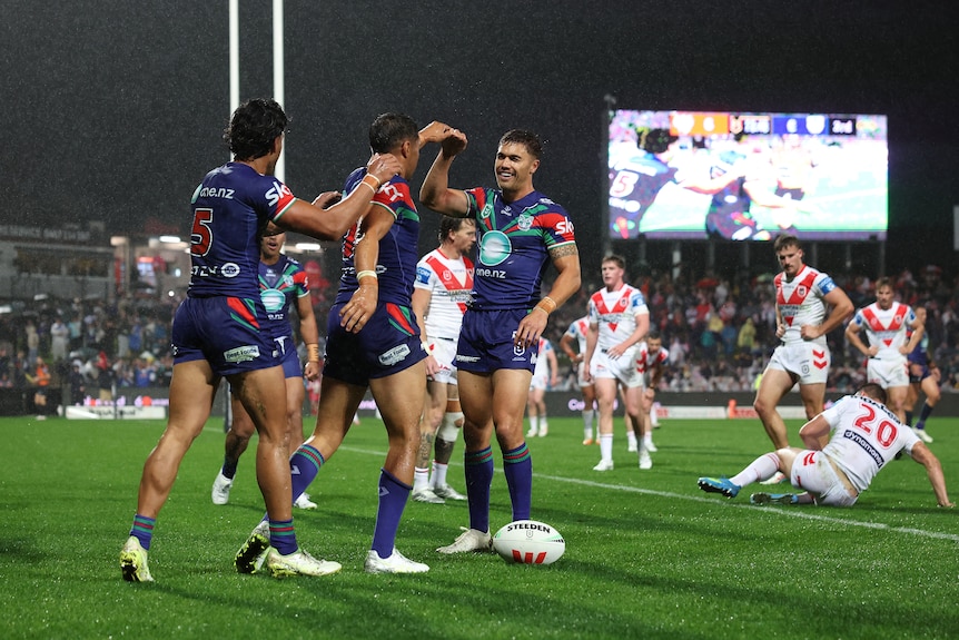 A group of rugby league players in blue celebrate a try with players in white behind them and on the ground