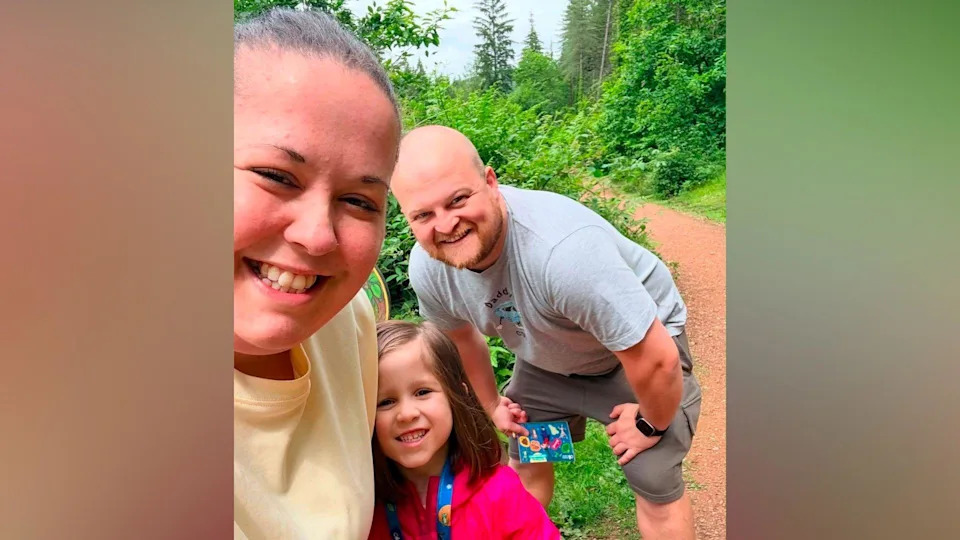 A family of three - a woman, a man and their daughter - smile as they stand on a bark path through a lush green forest. The man, who is bald, is wearing grey shorts and a grey T-shirt and is crouching with his hands resting on his knees while his daughter, who has shoulder-length brown hair, is wearing a pink coat. His wife has slicked-back brown hair and is wearing a yellow T-shirt.