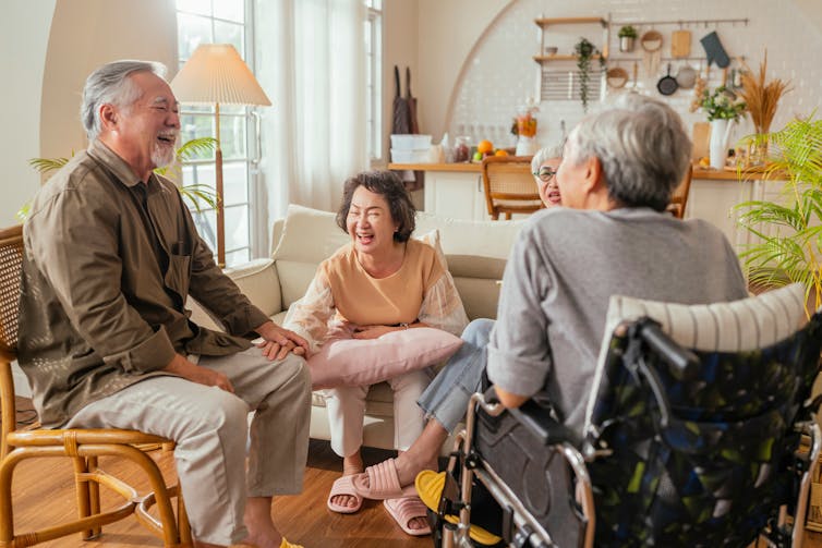 Four older adults laughing and sitting in a living room; one uses a wheelchair