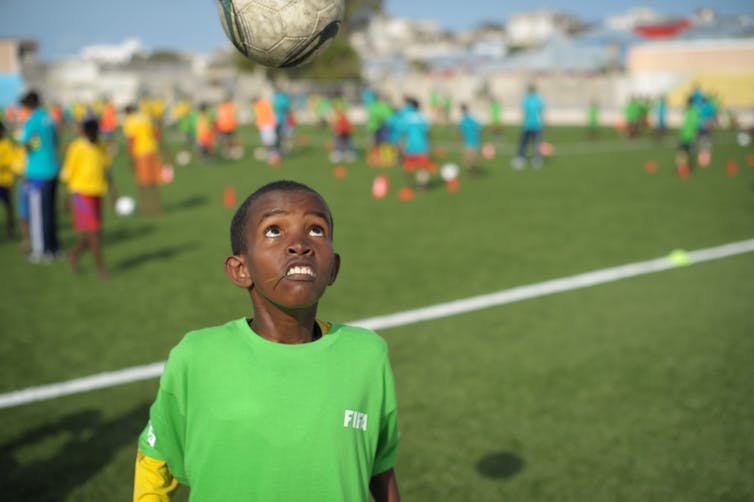 A young boy in a green football kit looks up at a ball in the air, on a football pitch.