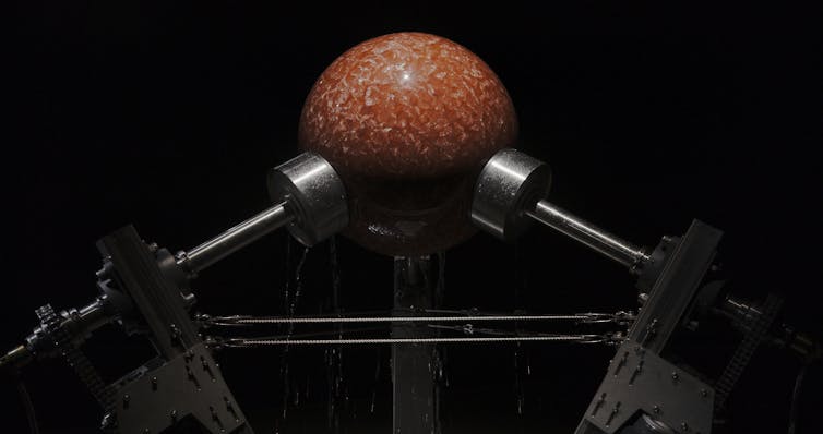 Ancient stromatolite fossil held between metal rollers in a mechanical framework. Water flows over the rock as the machine gradually grinds it into a smooth spherical shape.