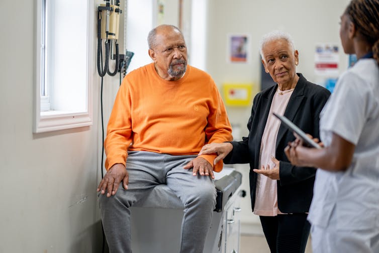 Older man sits on an exam table, older woman by his side, their doctor stands facing them.