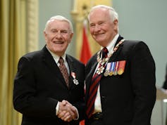Two white-haired men smile and shake hands.