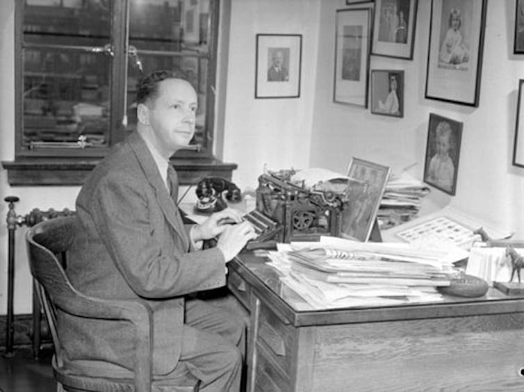 A black and white photo of a dark-haired man in a suit sitting at a desk.