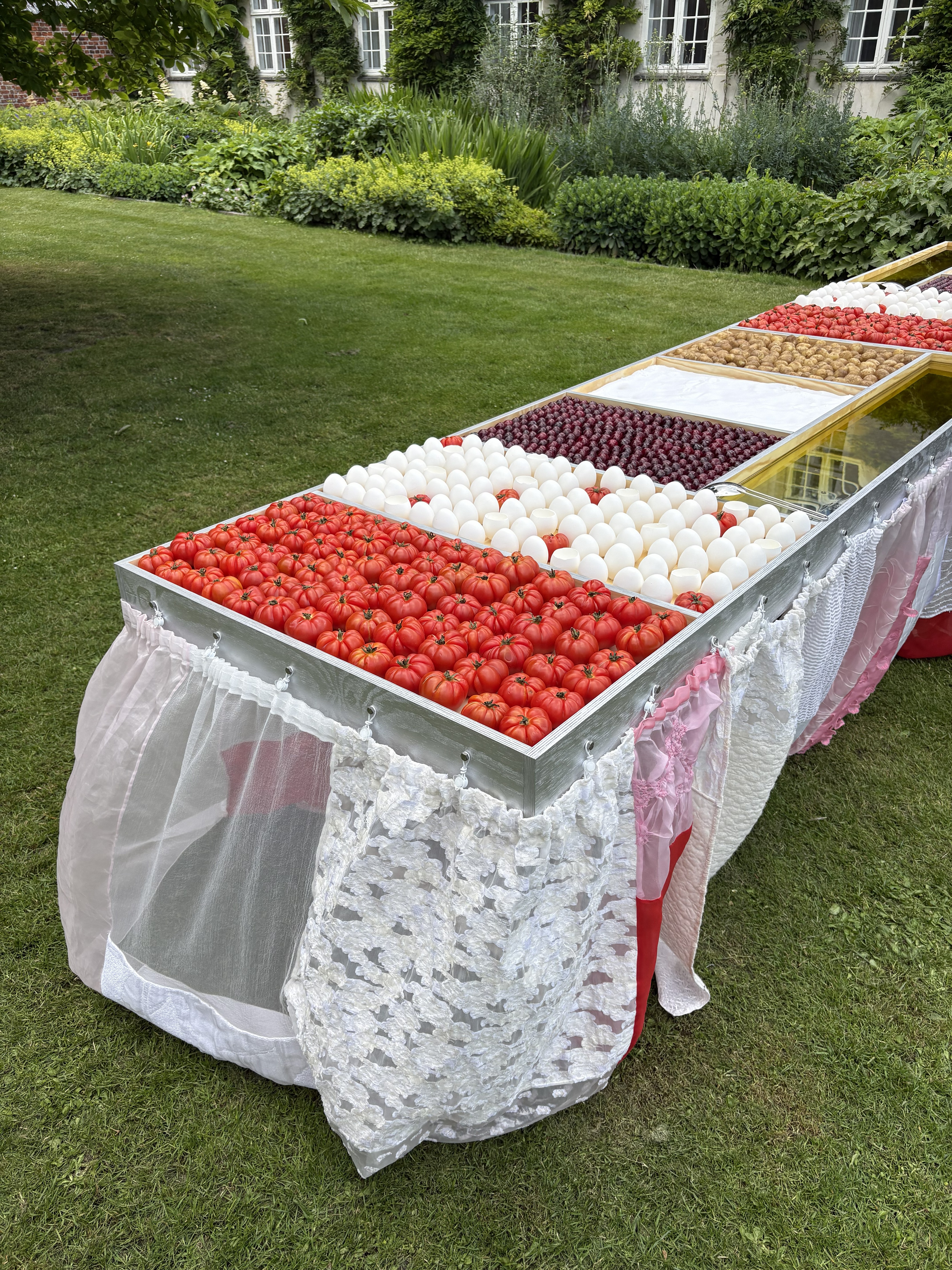 An outdoor table draped in different fabrics, and topped by a silver frame filled with tomatoes, boiled eggs, cherries, salt, potatoes, and olive oil against the grass.