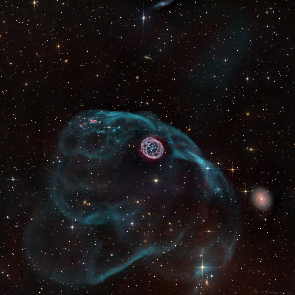 A gaseous structure that resembles a hamster wheel sits near the center of the image. Inside is a bright white dwarf star. There is a larger asymmetric gas bubble surrounding the inner nebula. A galaxy sits to the bottom right. The background is composed of foreground and background stars, as well as distant galaxies.