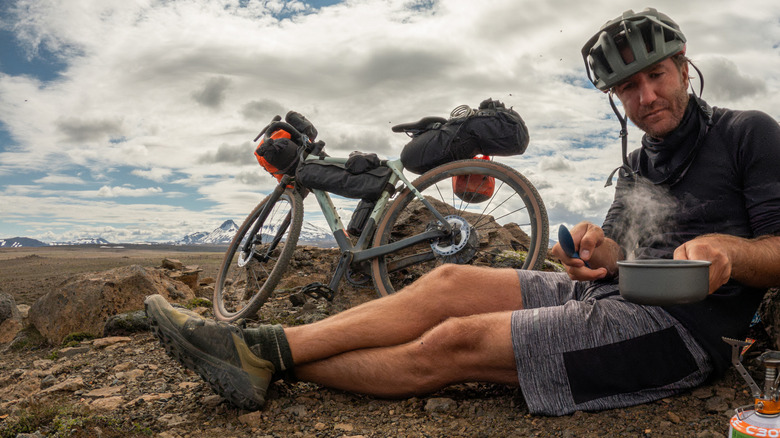 A bikepacker heats up his dinner in a pot next to his loaded bicycle on a remote plateau