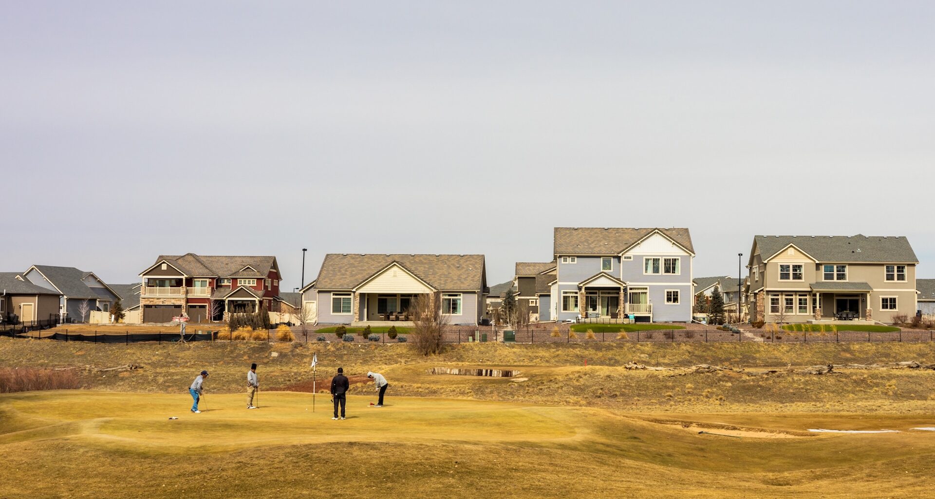 Four golfers practicing on a green, with suburban homes in the background under a clear sky.