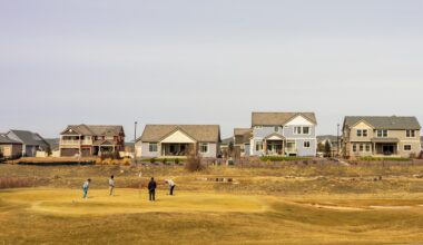 Four golfers practicing on a green, with suburban homes in the background under a clear sky.