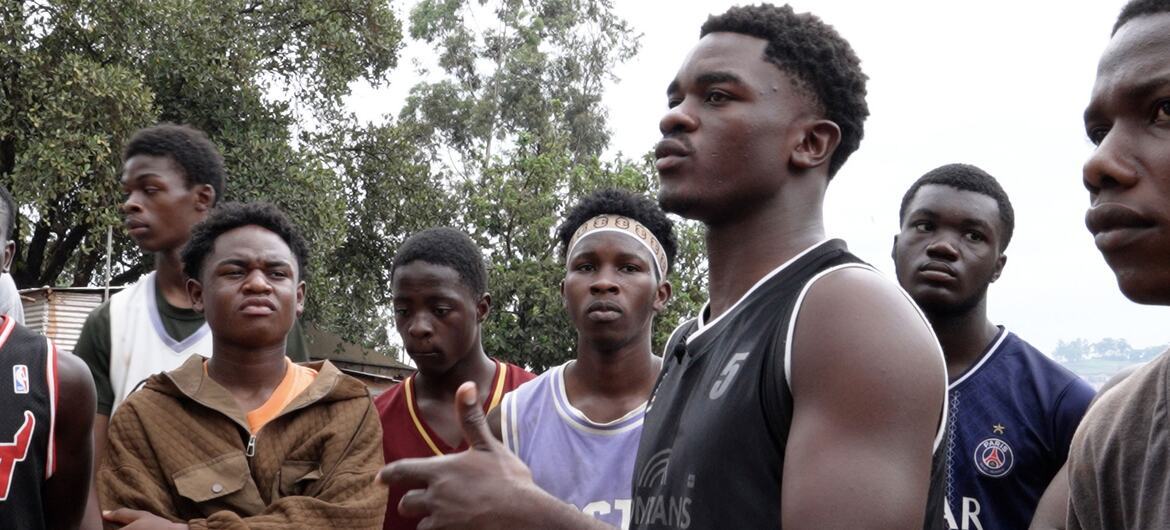 A group of young men, appearing to be refugees from the DRC, are gathered outdoors in Uganda. One man in a black jersey is speaking and gesturing to the others.