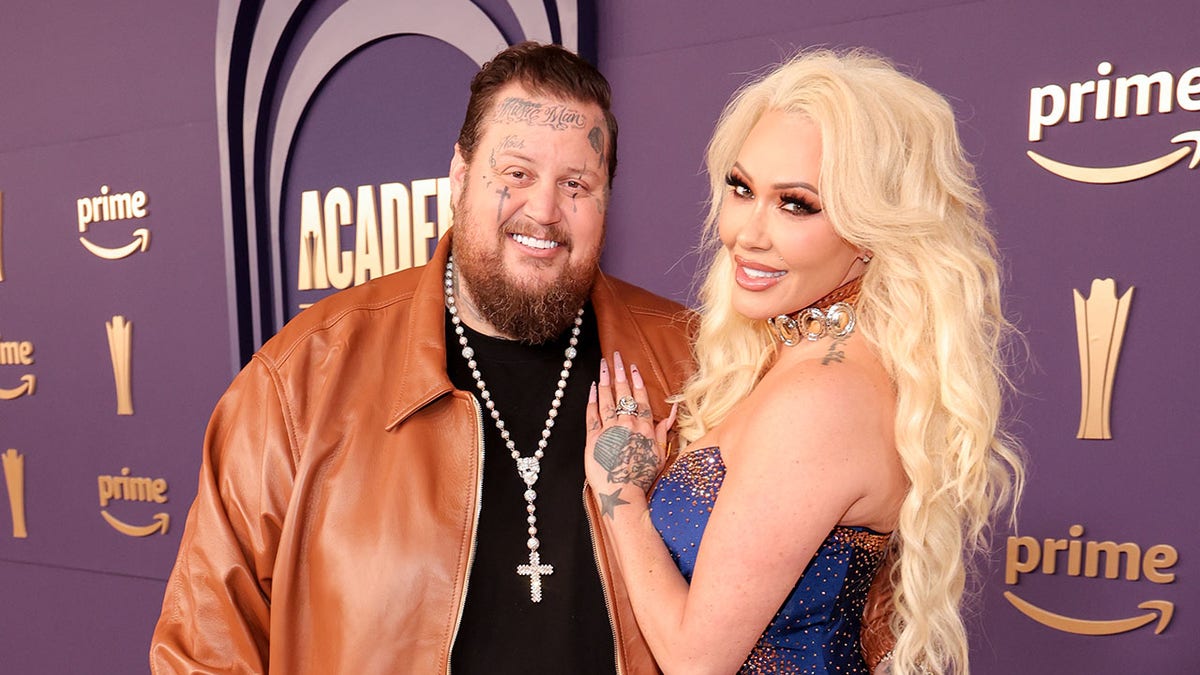 Jelly Roll smiling in a burnt orange jacket with Bunnie XO in a blue dress on the red carpet