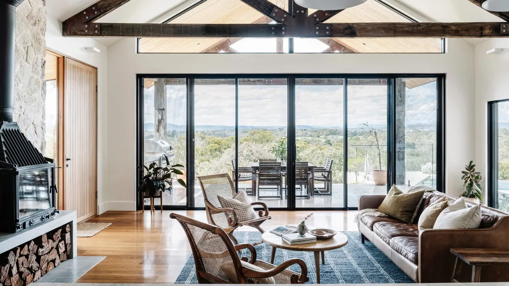 Barn style living room in a country style home with wooden beams and large windows that look out to a balcony.