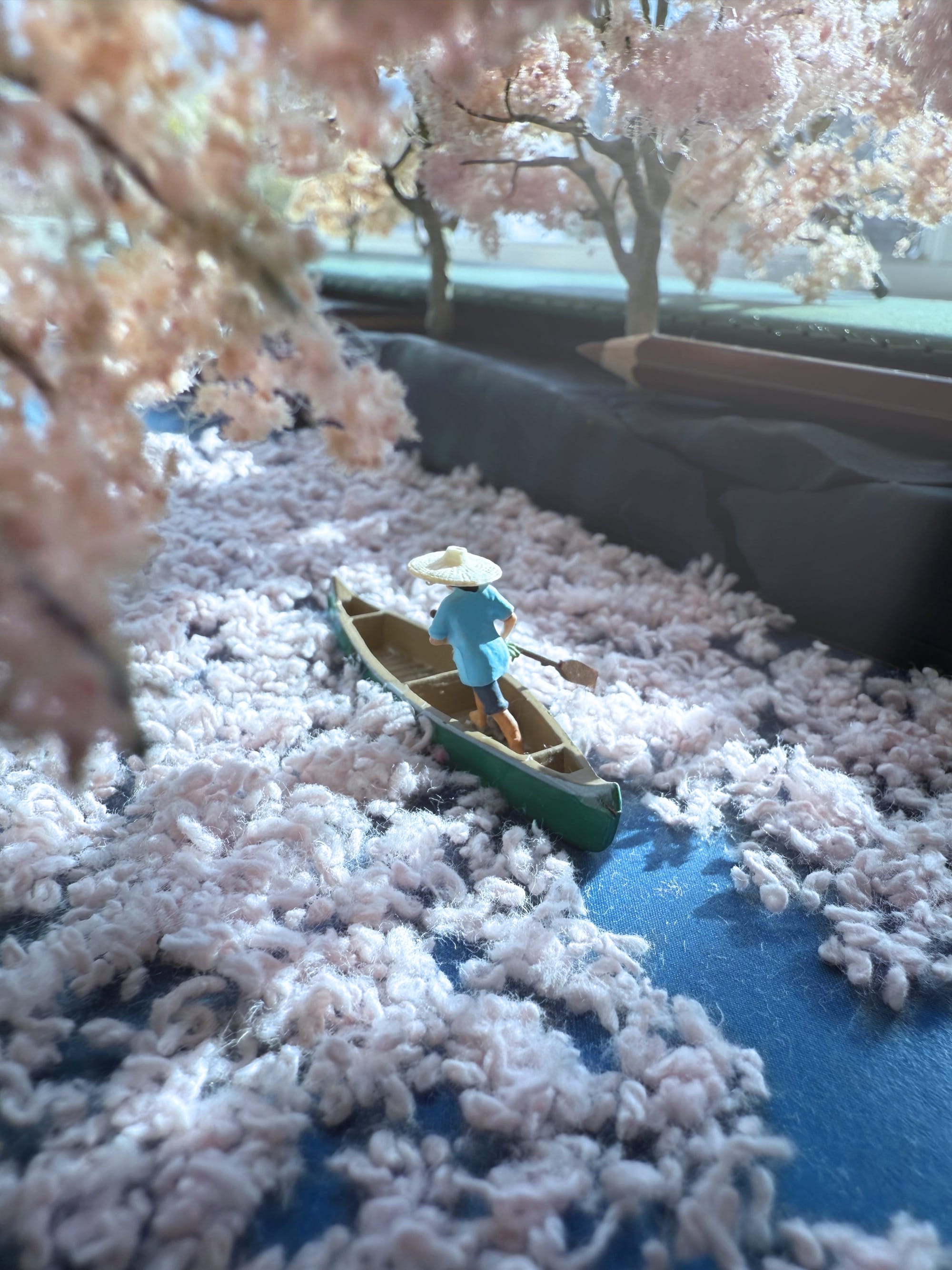 a photograph of a miniature tableau by Derrick Lin of a figure in a canoe, paddling down a river that is coated in dropped blossoms