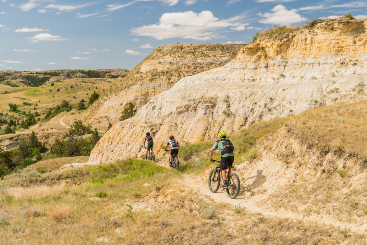Four mountain bikers riding along a trail beside a rocky hillside, with green vegetation and rolling hills in the background under a partly cloudy sky.