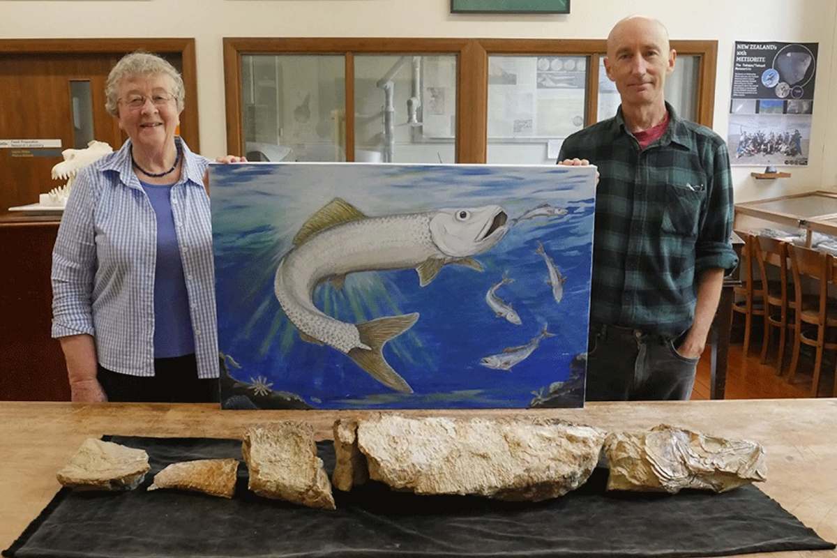 A woman and a man stand beside a painting of a fish, with fossilized fish remains displayed on the table in front of them.