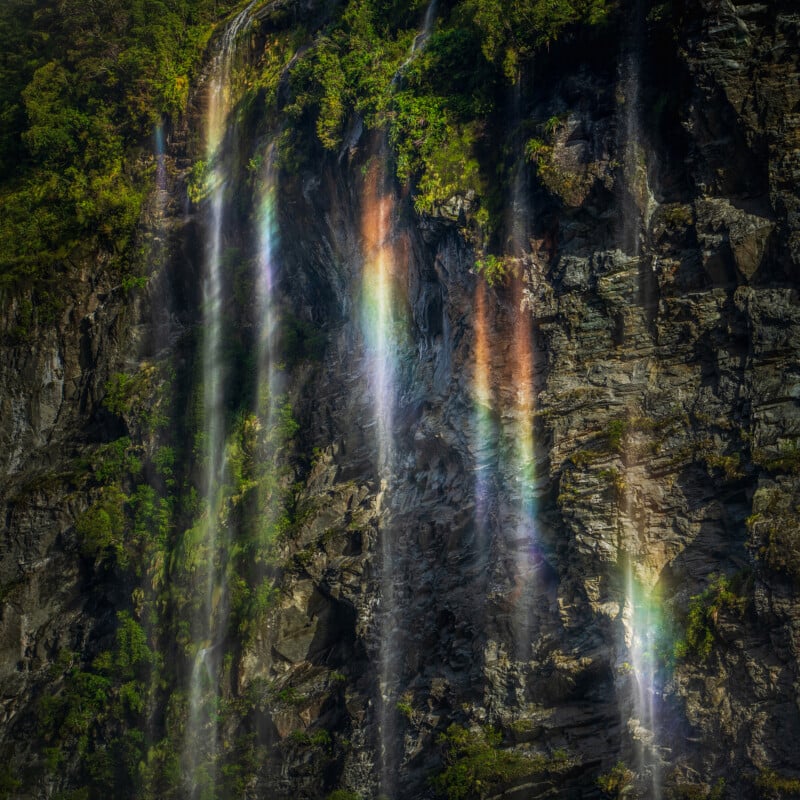Thin streams of waterfall cascade down a rocky cliff, illuminated by sunlight that creates visible rainbow colors. Lush green foliage grows along the top and sides of the cliff.