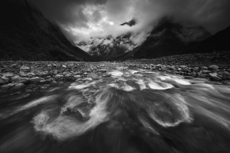 A dramatic black and white photo of a fast-flowing mountain river with rocks, surrounded by misty, cloud-covered peaks beneath a moody, overcast sky.