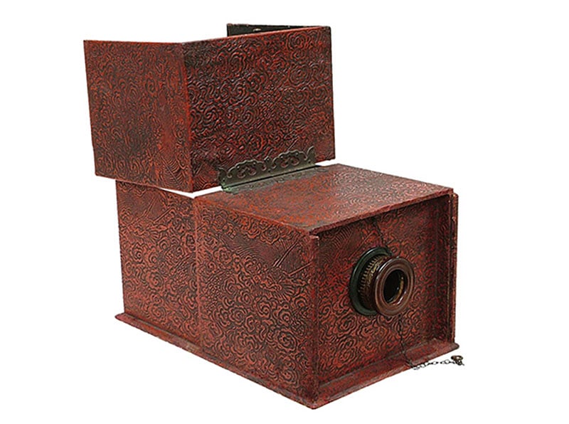 An antique, ornately patterned red camera obscura box with a lens attached at the front and a decorative metal latch on the side, used historically for projecting images.