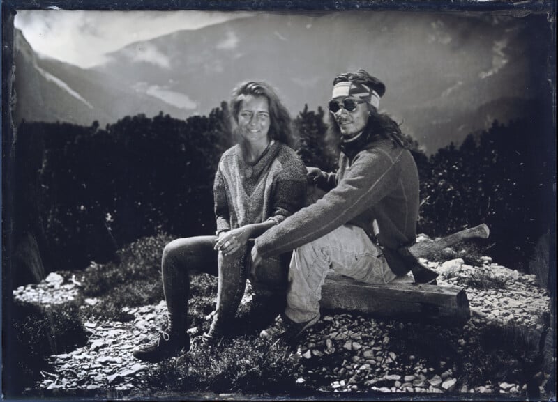 Two people sit outdoors on a log, smiling at the camera, with mountains and trees in the background. One person wears sunglasses and a headband; the other wears a sweater and has loose hair. The image is black and white.