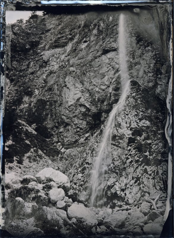 A thin waterfall cascades down a rocky cliff face in a black and white photograph with a vintage feel, surrounded by rocks and foliage at its base.