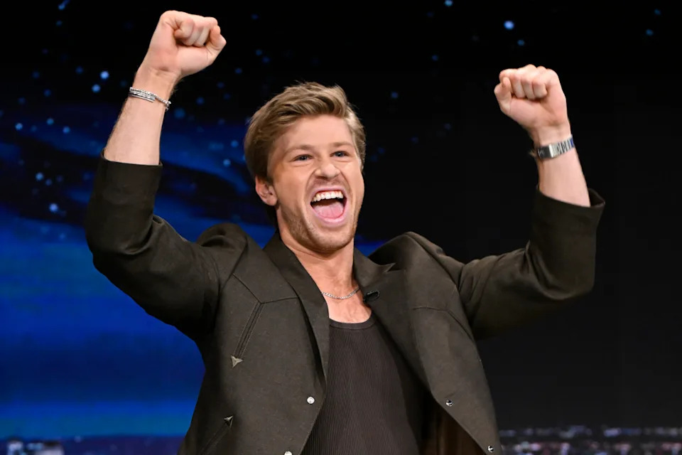 Robert Irwin in a black suit jacket, smiling with his hands above his head in celebration.